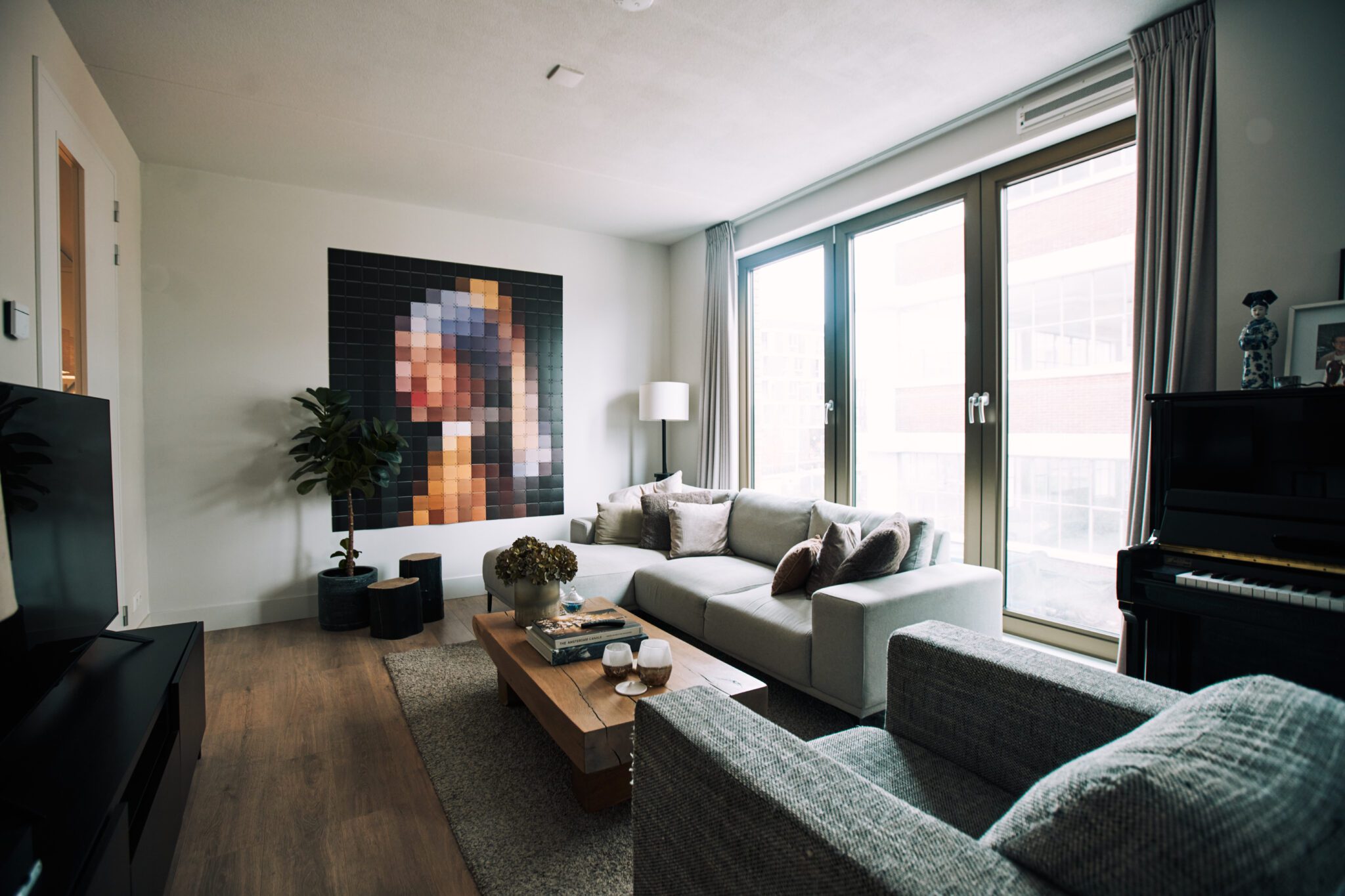 Modern ingerichte woonkamer met grote ramen, een lichtgrijze hoekbank, houten salontafel en een opvallend pixelkunstwerk van 'Meisje met de parel' aan de muur. Rechts in beeld een zwarte piano met decoratieve items.