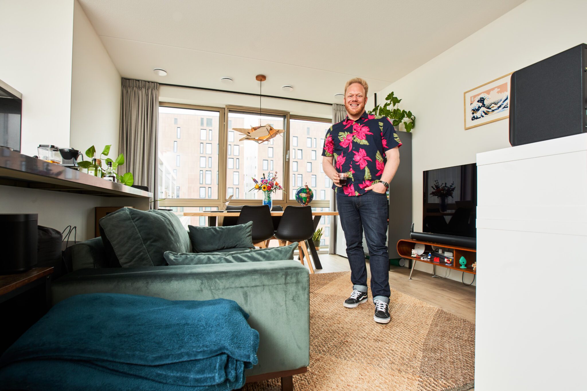 Rob Thijs staat ontspannen met een drankje in zijn hand in zijn kleurrijke woonkamer, met achter hem een designkast, flatscreen-tv en een boekenkast vol boeken en persoonlijke items.
