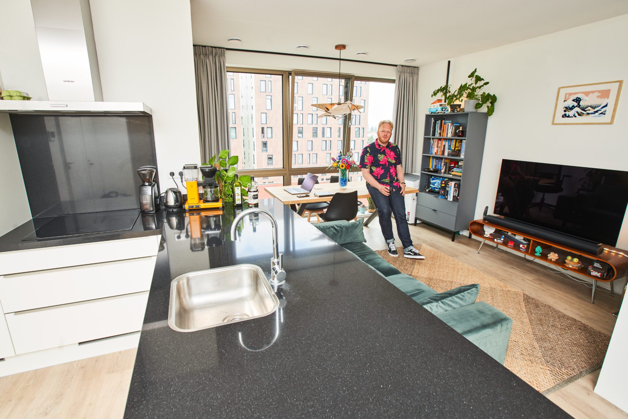 Rob Thijs staat ontspannen met een drankje in zijn hand in zijn kleurrijke woonkamer, met achter hem een designkast, flatscreen-tv en een boekenkast vol boeken en persoonlijke items.
