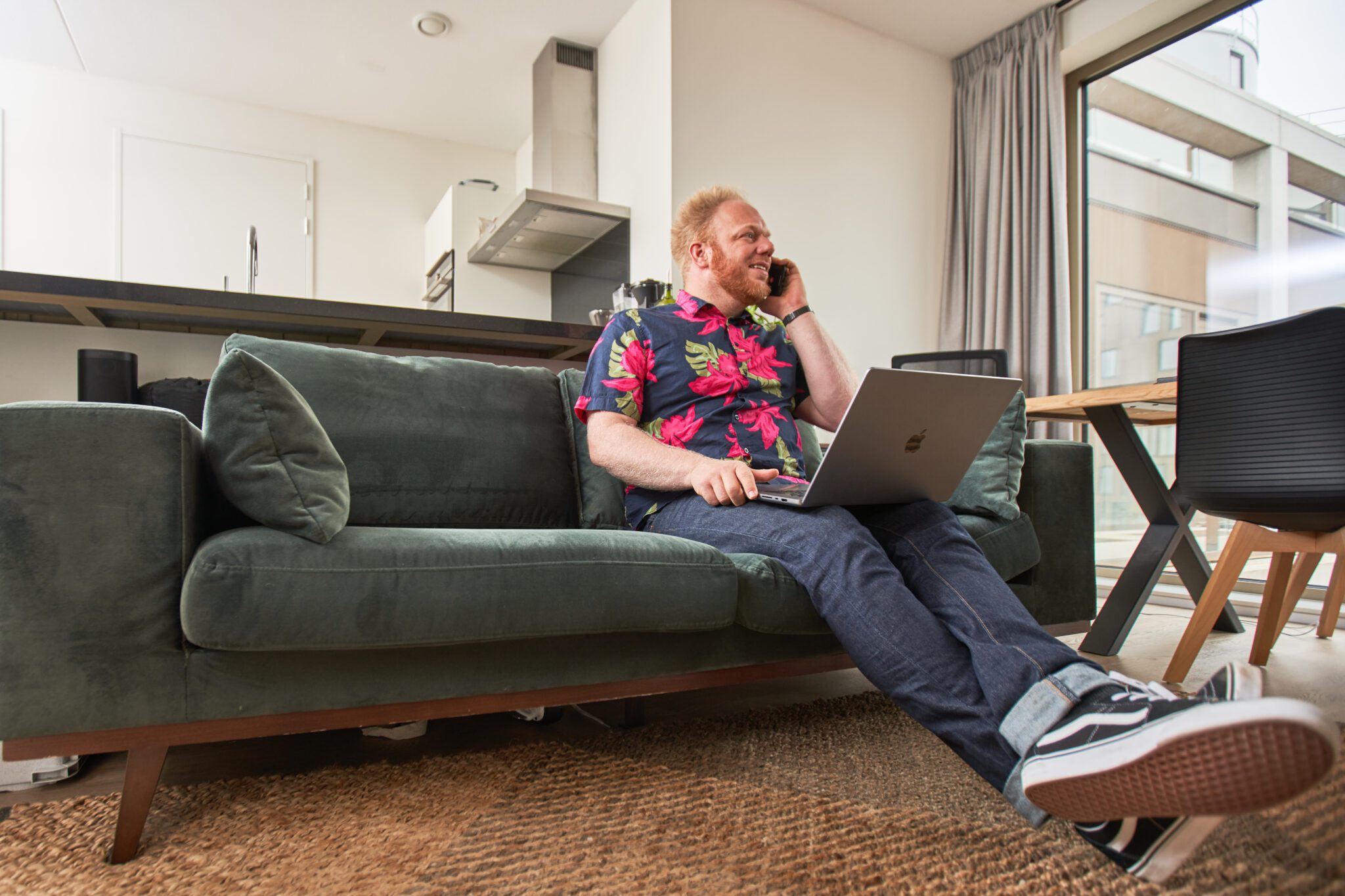 Rob zit relaxed op een groene fluwelen bank, met zijn laptop op schoot en een telefoon aan zijn oor, in gesprek terwijl hij werkt in zijn woonkamer.