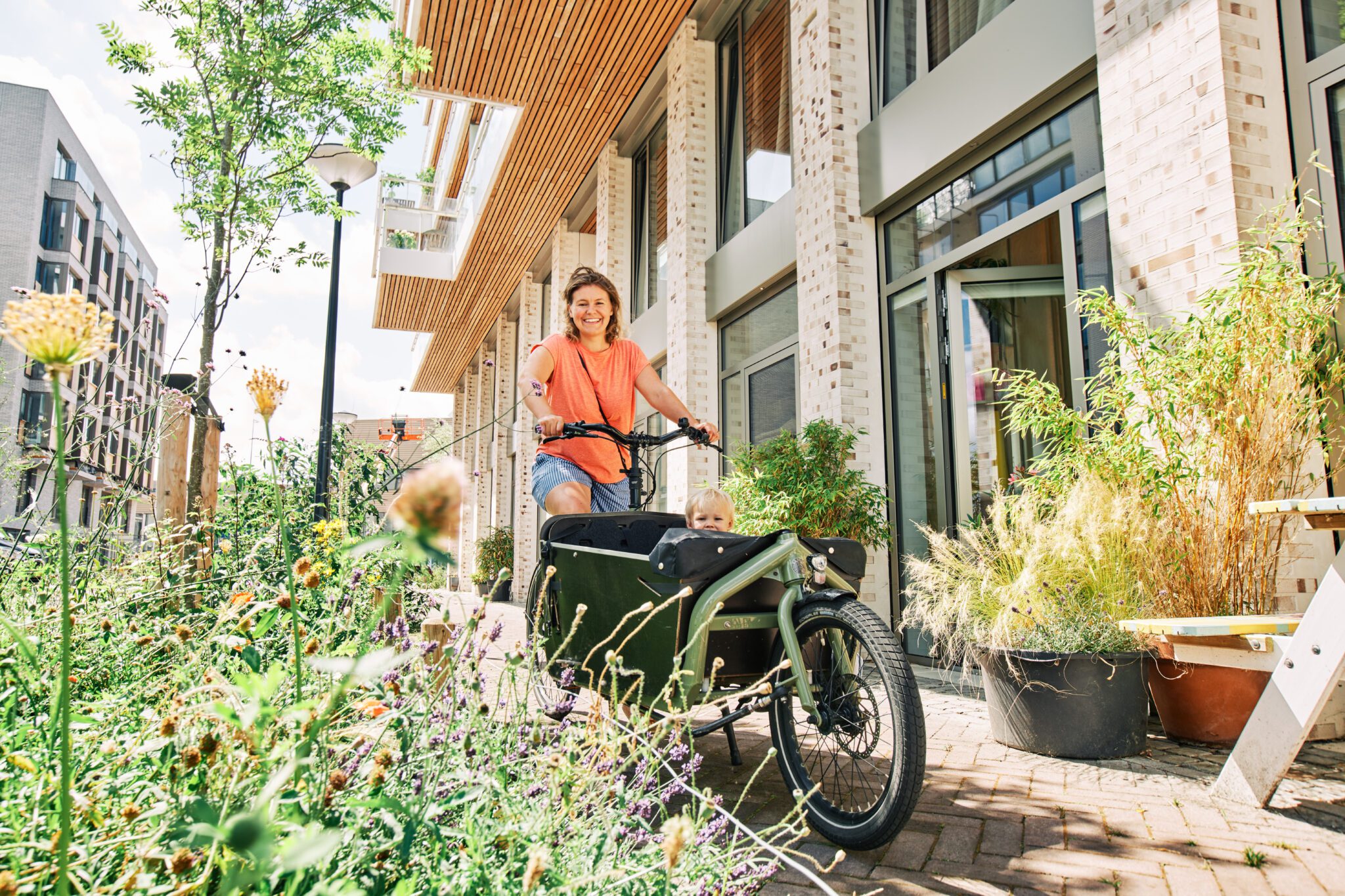 Vrouw op een bakfiets met jong kind in het zitgedeelte, fietst lachend langs een bloemrijke geveltuin in een moderne straat in Cruquius, op een zonnige dag.
