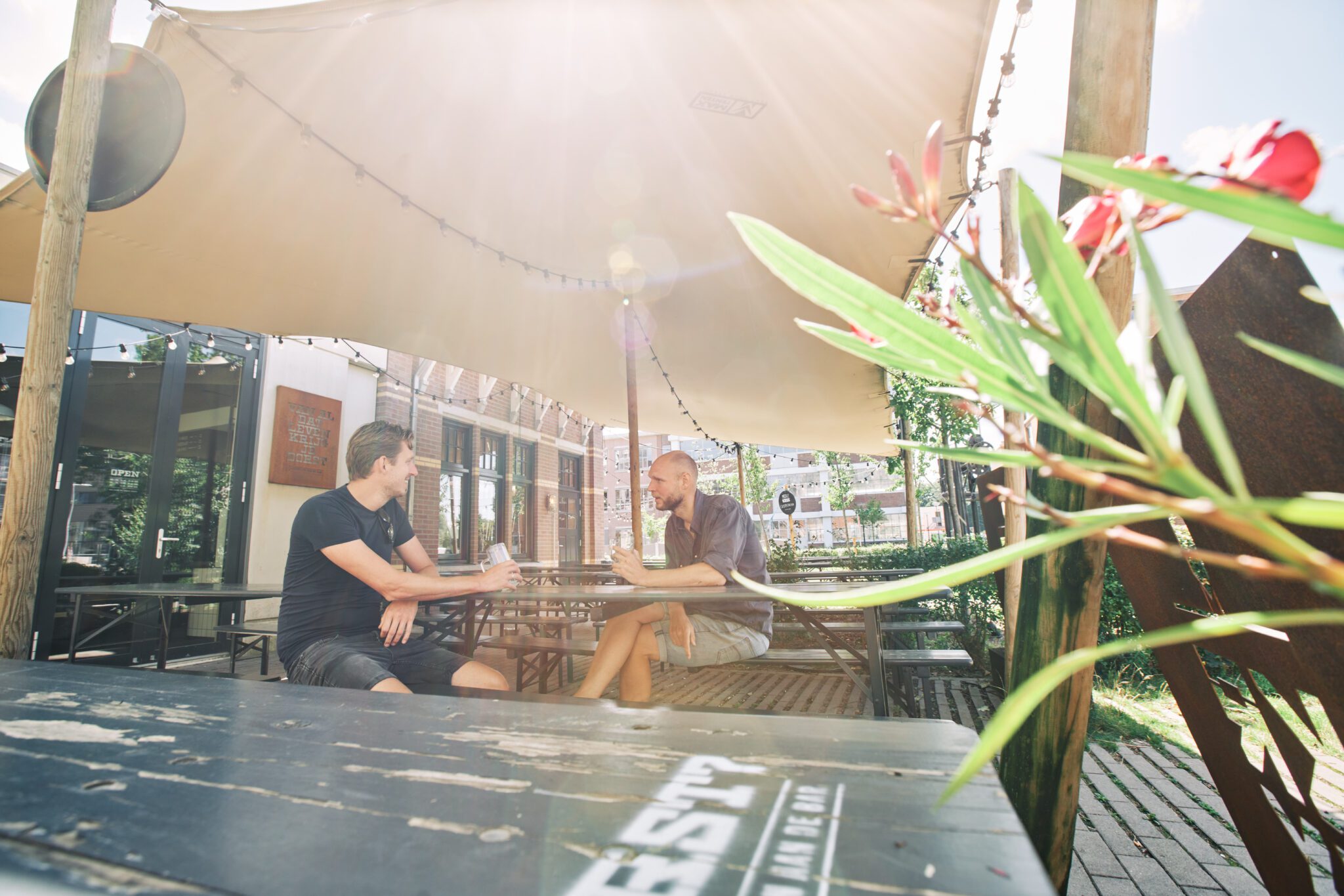 Twee mannen zitten in de zon op het terras van brouwerij KRUX onder een schaduwdoek, met houten picknickbanken, string lights en zomerse planten op de voorgrond.