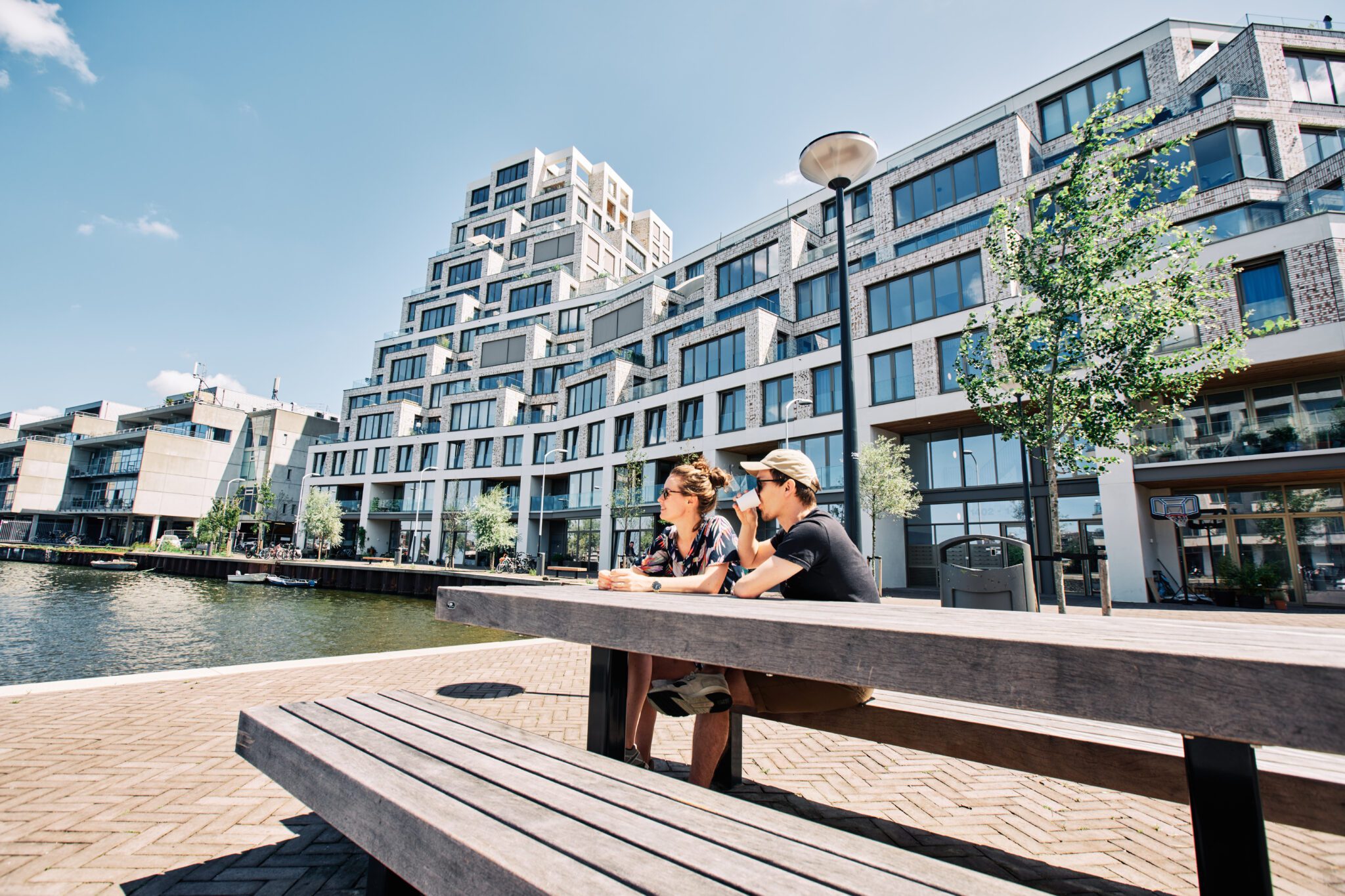 Twee mensen zitten op een bankje aan het water in Cruquius, met op de achtergrond het iconische trappengebouw en moderne appartementengebouwen onder een heldere blauwe lucht.