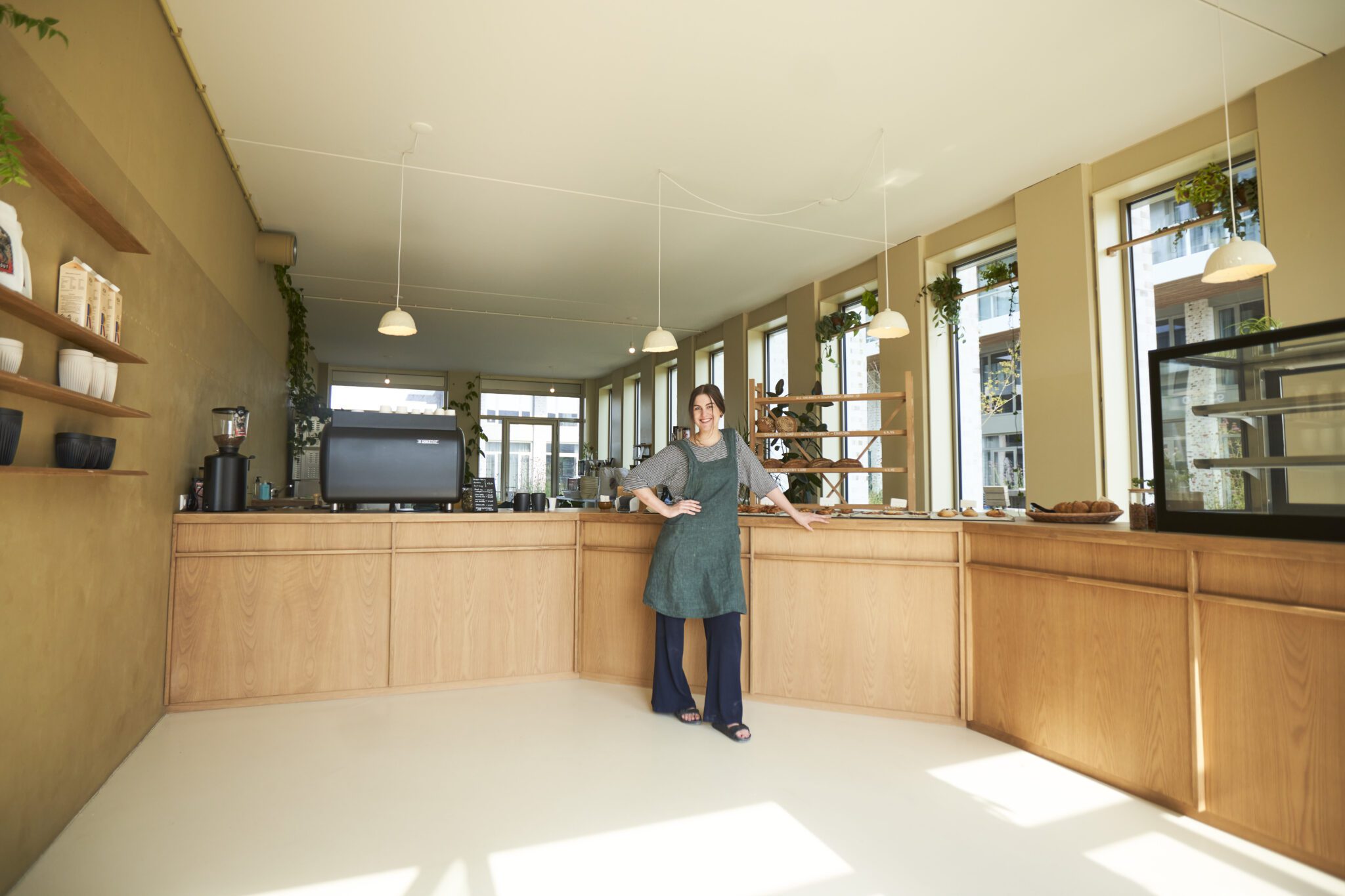 Ondernemer van Millie Vanillie poseert trots in een strakke, minimalistische bakkerij met houten toonbank en rekken vol daglicht.
