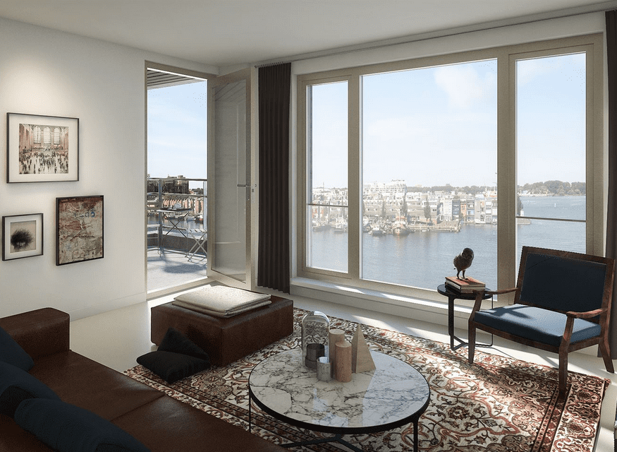 Zitkamer met uitzicht over het water, grote ramen, klassieke vloerkleed, marmeren salontafel en toegang tot een balkon met zitgelegenheid.