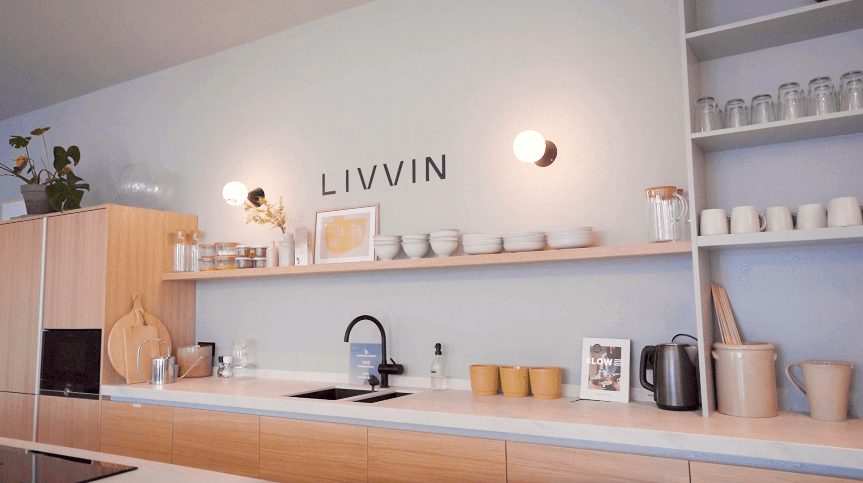 Moderne en minimalistische keuken in de LIVVIN community space, met houten kastfronten, open planken met servies, zwarte kraan en de merknaam 'LIVVIN' op de muur.