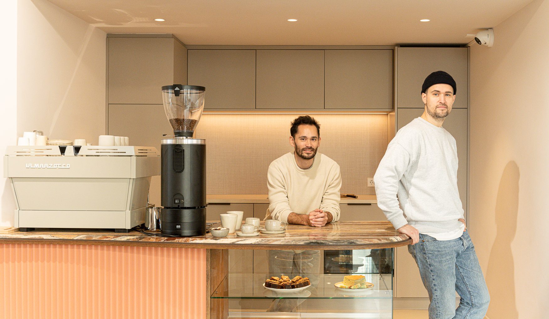 Ondernemers van Joki poseren in een stijlvolle koffiezaak achter de bar met een La Marzocco machine en gebakjes.