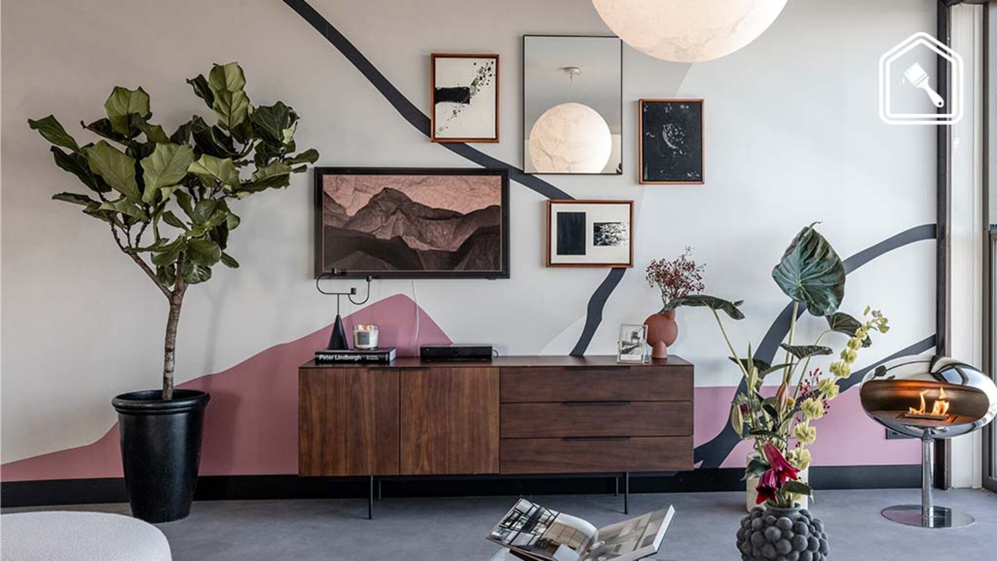 Moderne woonkamer met een houten dressoir, grote kamerplant, wandkunst en een designhaard. De muur is beschilderd met grafische lijnen in roze, grijs en zwart, en afgewerkt met decoratieve objecten en kunstboeken.