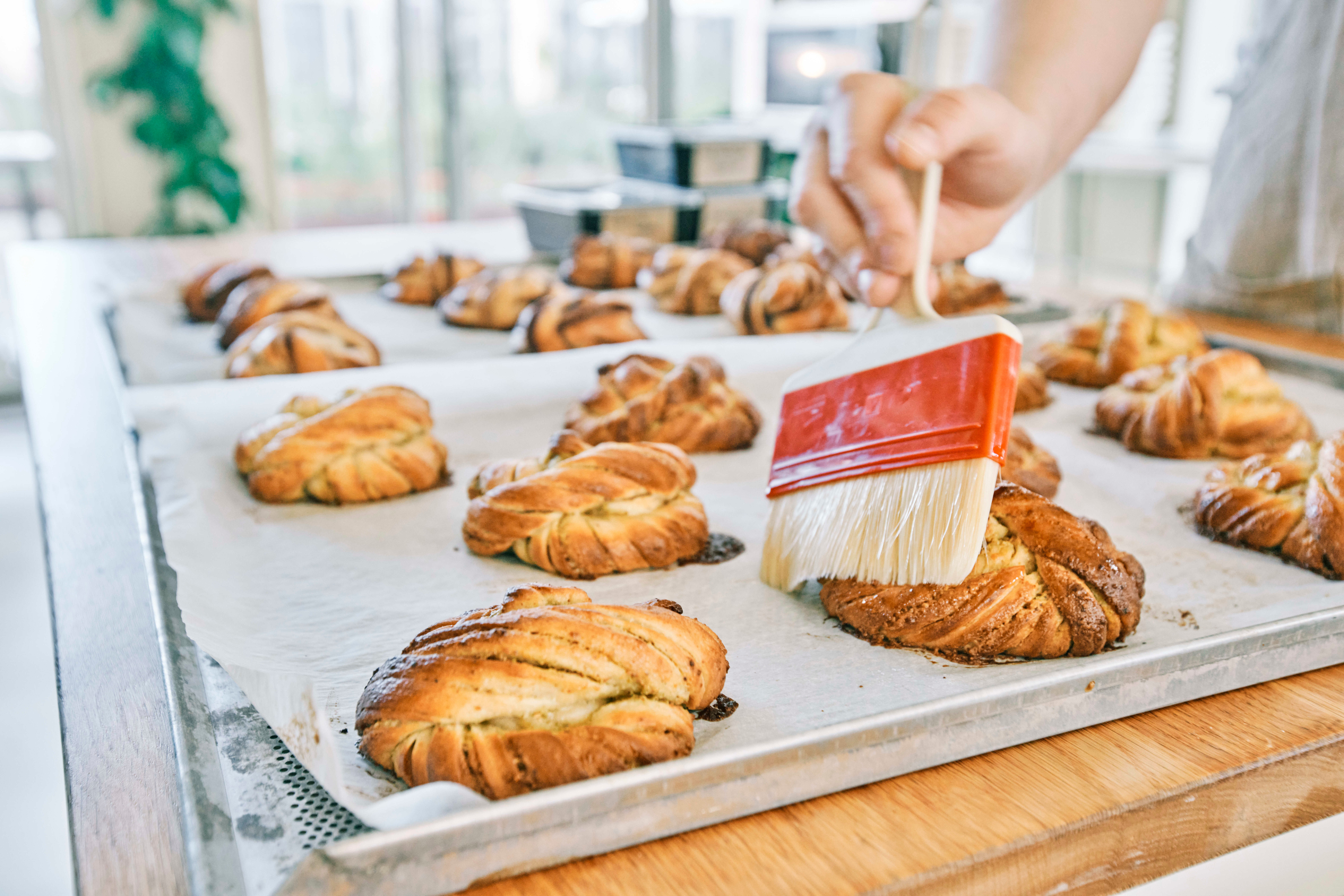 Close-up van versgebakken kaneelknoopsbroodjes op een bakplaat, terwijl een hand een van de broodjes afwerkt met een glazuurkwast in een lichte, ambachtelijke bakkerijomgeving.