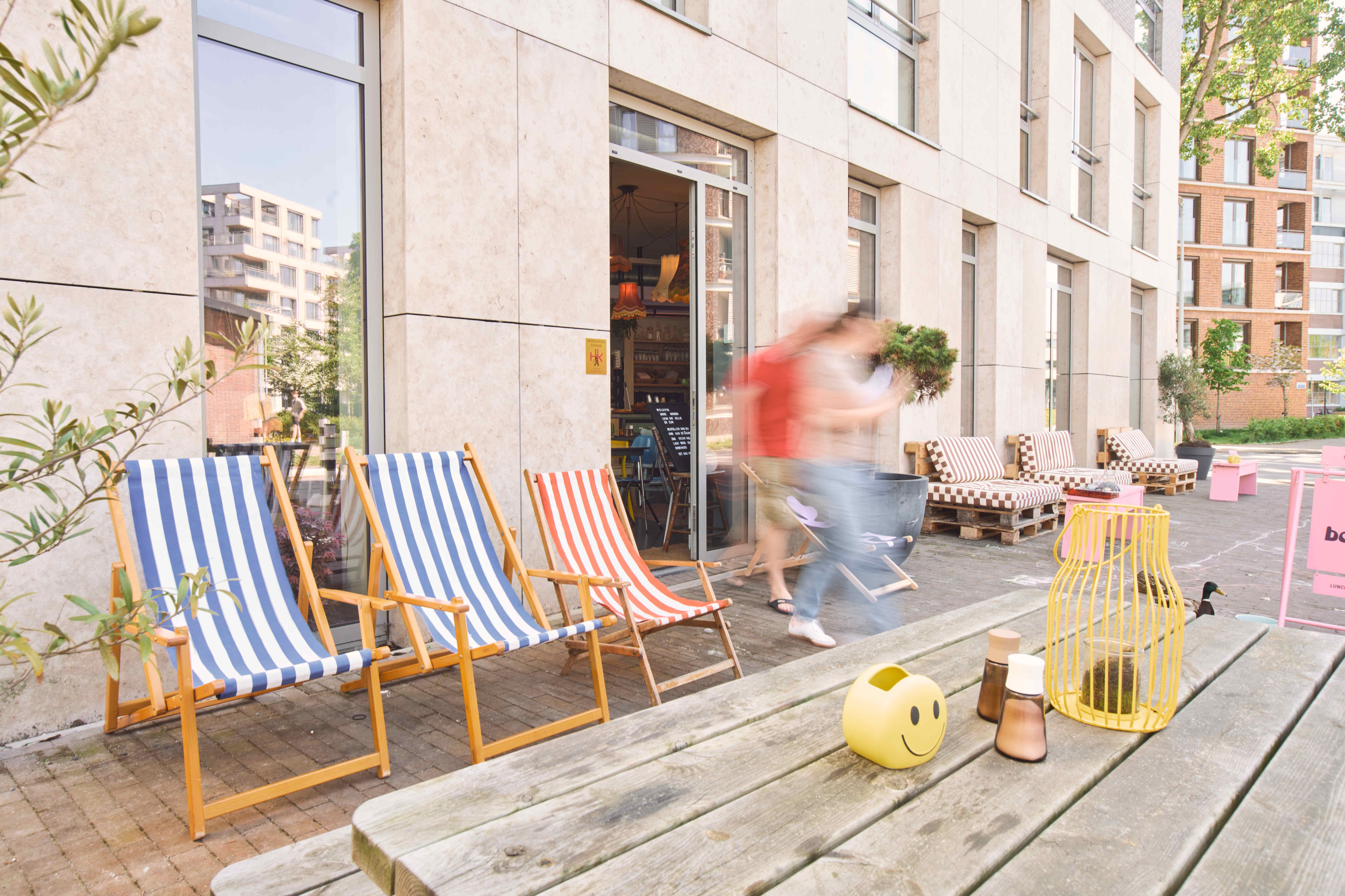 Kleurrijk terras van een lokale horecazaak in Cruquius met gestreepte ligstoelen, houten tuinmeubilair en speelse decoratie; twee mensen lopen langs in zomerse sfeer.
