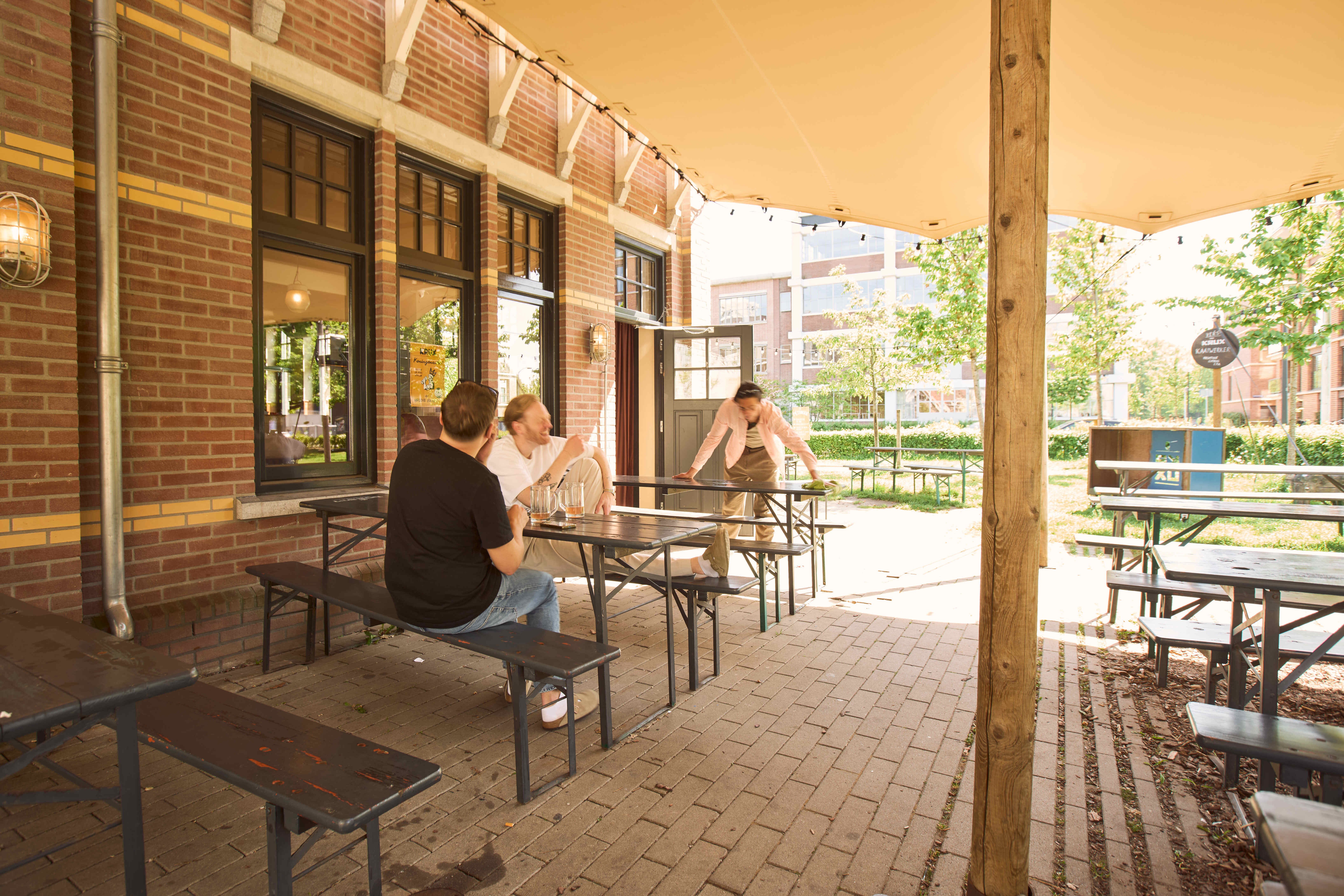 Twee mannen zitten in de zon op het terras van brouwerij KRUX onder een schaduwdoek, met houten picknickbanken en string lights.
