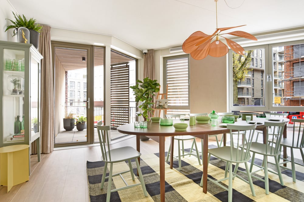 Lichte en moderne eetkamer in 2peer met grote ramen, ronde houten eettafel, mintgroene stoelen en een speelse hanglamp in bladvorm. De ruimte is stijlvol ingericht met groene accenten en biedt toegang tot een balkon met plantenbakken.